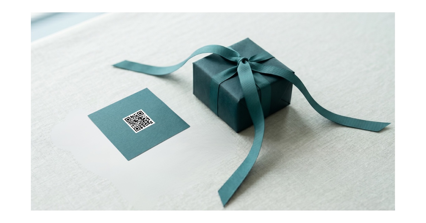A small wrapped gift box with a teal ribbon sits on a textured tabletop next to a teal QR-code card.